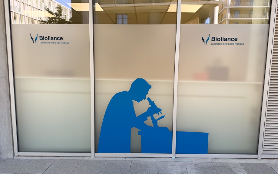 Vitrophanie sur mesure pour laboratoire d'un adhésif opaque à dos gris sur façade vitrée, silhouette d'un technicien au microscope